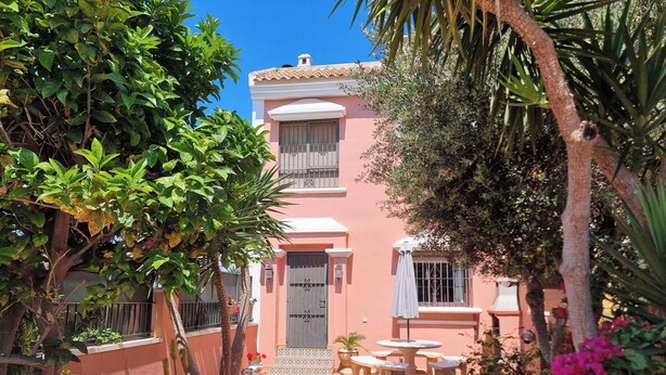A courtyard with a tiled floor, surrounded by a pink building with a balcony and a door. There are potted plants and a small table with chairs. - Three double bedroom townhouse in lovely village (San Javier, Murcia)