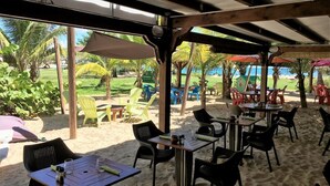 An outdoor seating area with tables and chairs, a covered structure, and a view of the beach and palm trees. - Ti Treasure Apartment Ground Floor T3 Crystal Beach (Saint-François)