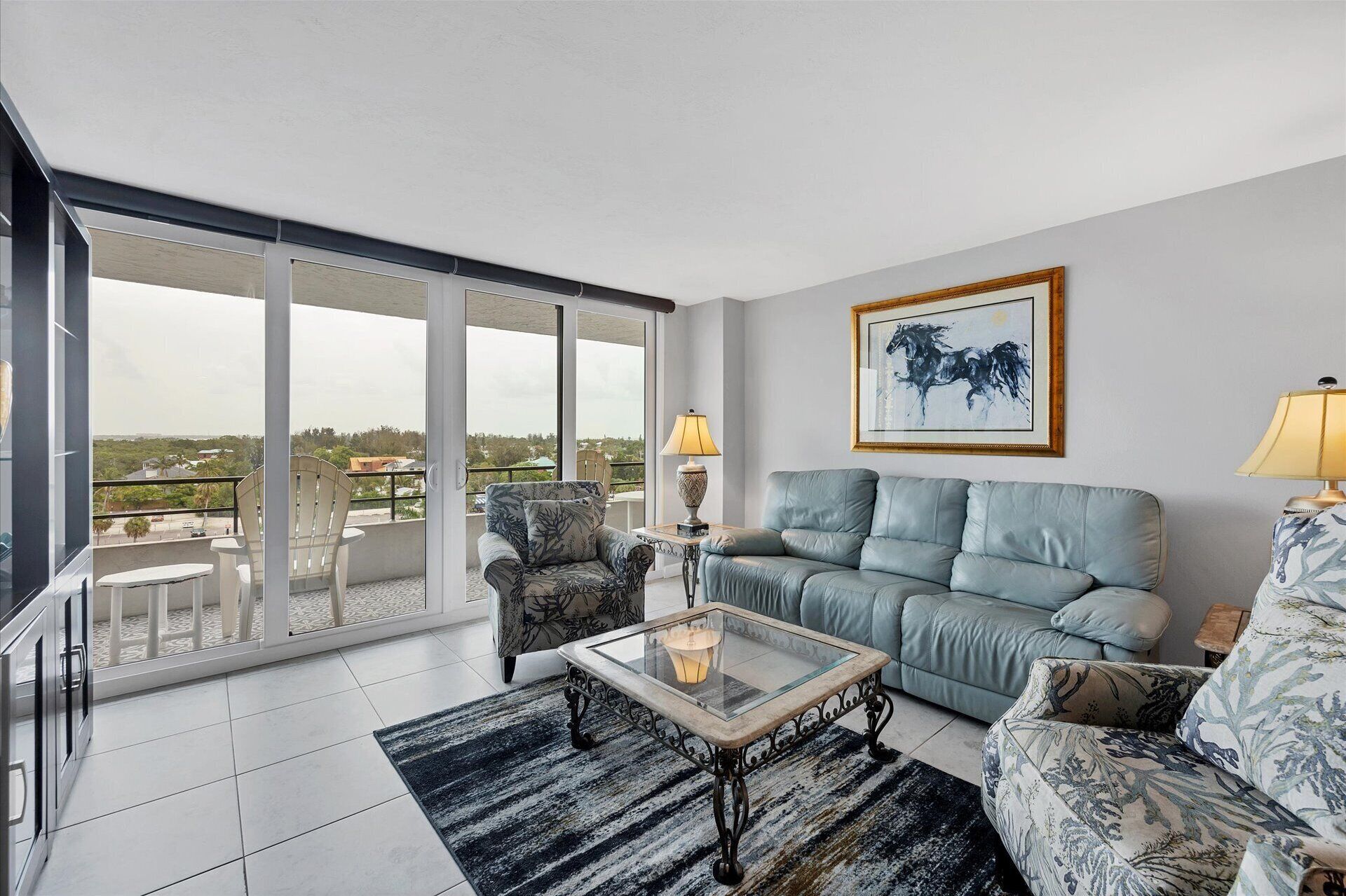 A spacious living room with a large glass door leading to a balcony, a grey sofa, a coffee table, and a framed artwork on the wall.
