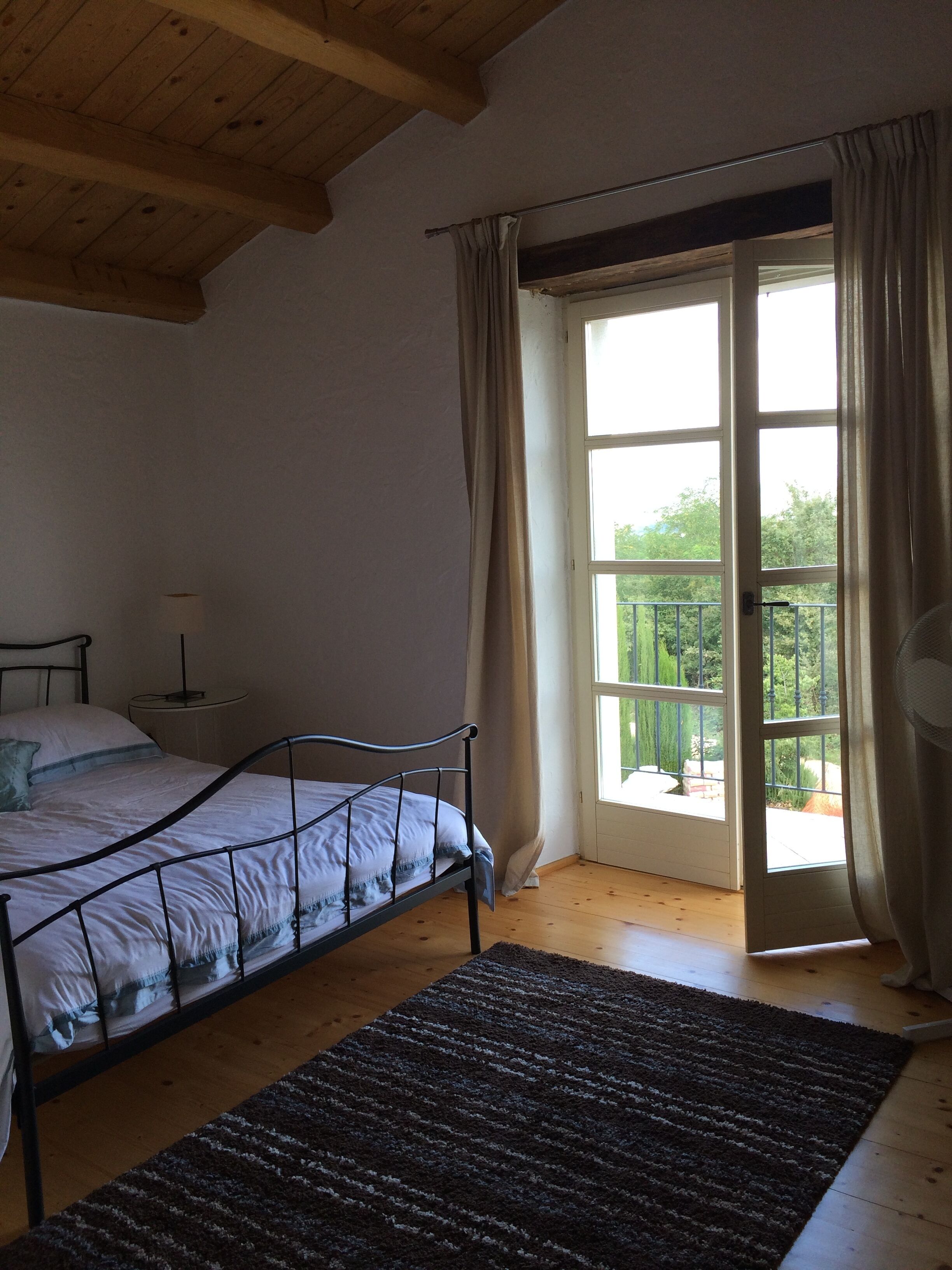 Un dormitorio con cama, mesita de noche, ventana con cortinas y vista a zonas verdes.