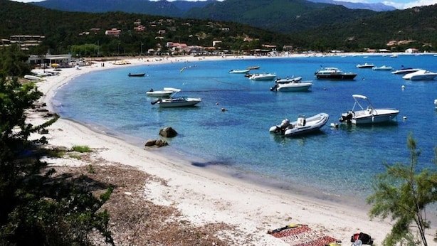 Une plage côtière bordée de bateaux, aux eaux cristallines, avec une chaîne de montagnes en arrière-plan.