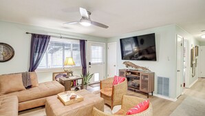 Uma sala de estar com sofá, puf, cadeiras de vime, uma mesa com bandeja, ventilador de teto e uma TV fixada na parede.