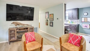Uma sala de estar com televisão de tela plana, um móvel de entretenimento de madeira, duas cadeiras de vime com almofadas vermelhas e uma cozinha visível através de uma porta aberta.