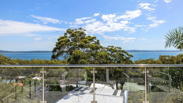 A balcony with a glass railing overlooking a coastal landscape with trees and a view of the sea. - Water Views on Wallawa - Gorgeous Nelson Bay Water Views (Nelson Bay)
