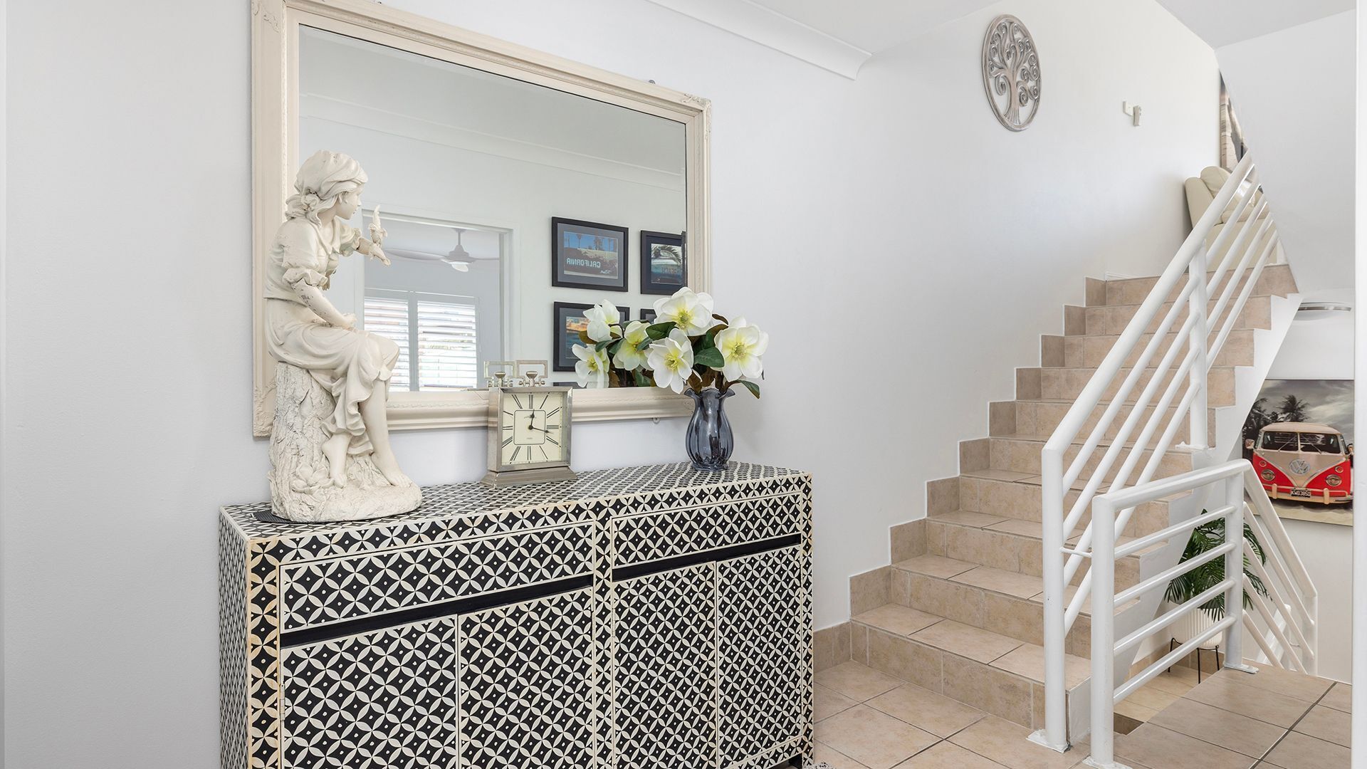 Eine Treppe mit weißen Geländern und beigen Fliesen, ein weißer Konsolentisch mit geometrischem Muster, eine Statue, eine Uhr und eine Vase mit Blumen.
