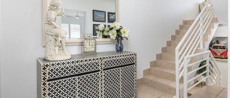 Eine Treppe mit weißen Geländern und beigen Fliesen, ein weißer Konsolentisch mit geometrischem Muster, eine Statue, eine Uhr und eine Vase mit Blumen.