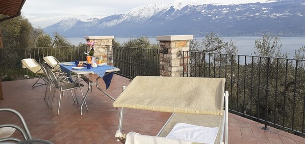 A terrace with a table set for dining, chairs, and a sunshade. A stone pillar and railing are visible.