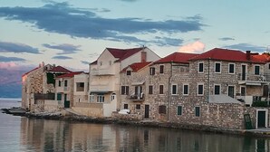 A row of stone buildings with red roofs by a calm body of water, reflecting the structures and sky. - Beachfront apt. with big terrace &  garden (Postira)