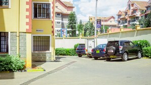 A residential building with a yellow facade, a gated entrance, and a parking area with two cars. - Lomu Homes Kilimani (Nairobi)