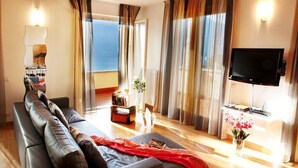 A modern living room with a large sofa, a flat-screen TV, and a view of the sea through large windows.