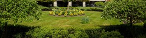 Stroll through the serene garden, surrounded by vibrant flowers and lush greenery, with trees providing shade and a low wall enclosing the peaceful space.
