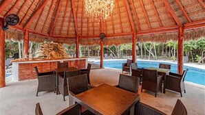 Un espace extérieur aménagé pour les repas, avec un toit de chaume, des meubles en osier et une piscine en arrière-plan.