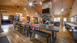 A spacious wooden cabin interior with a large dining table, benches, a stone fireplace, and a kitchen area.