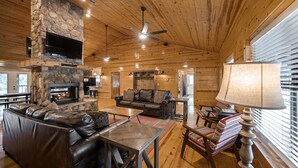 A log cabin interior with a stone fireplace, leather sofa, and a television mounted on a stone wall.