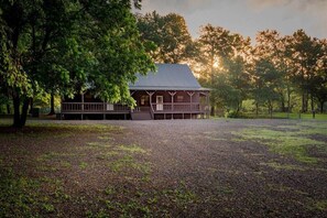 Une cabane en bois rustique, dotée d’un porche, entourée d’une zone recouverte de gravier et d’arbres.