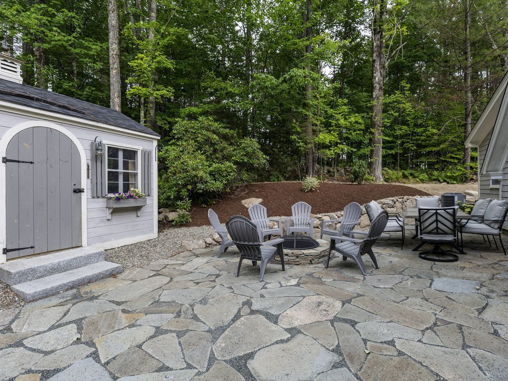 A stone patio with Adirondack chairs and a fire pit, surrounded by a wooded area.