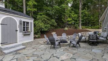 A stone patio with Adirondack chairs and a fire pit, surrounded by a wooded area.