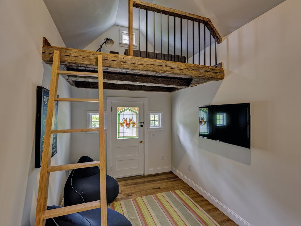 Un dormitorio tipo loft con una escalera de madera, una silla negra, un tapete a rayas, un televisor en la pared y una puerta con un vitral.