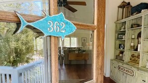 Une porte en bois avec un numéro, une rambarde blanche et une vue sur une pièce dotée d’un plancher en bois et d’un meuble sur lequel est inscrit « MAISON DE PLAGE ».
