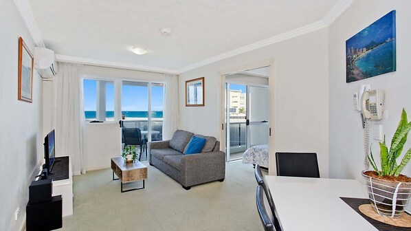 Een moderne woonkamer met een grijze bank, een salontafel en een televisie. Er is een balkon met uitzicht op zee.