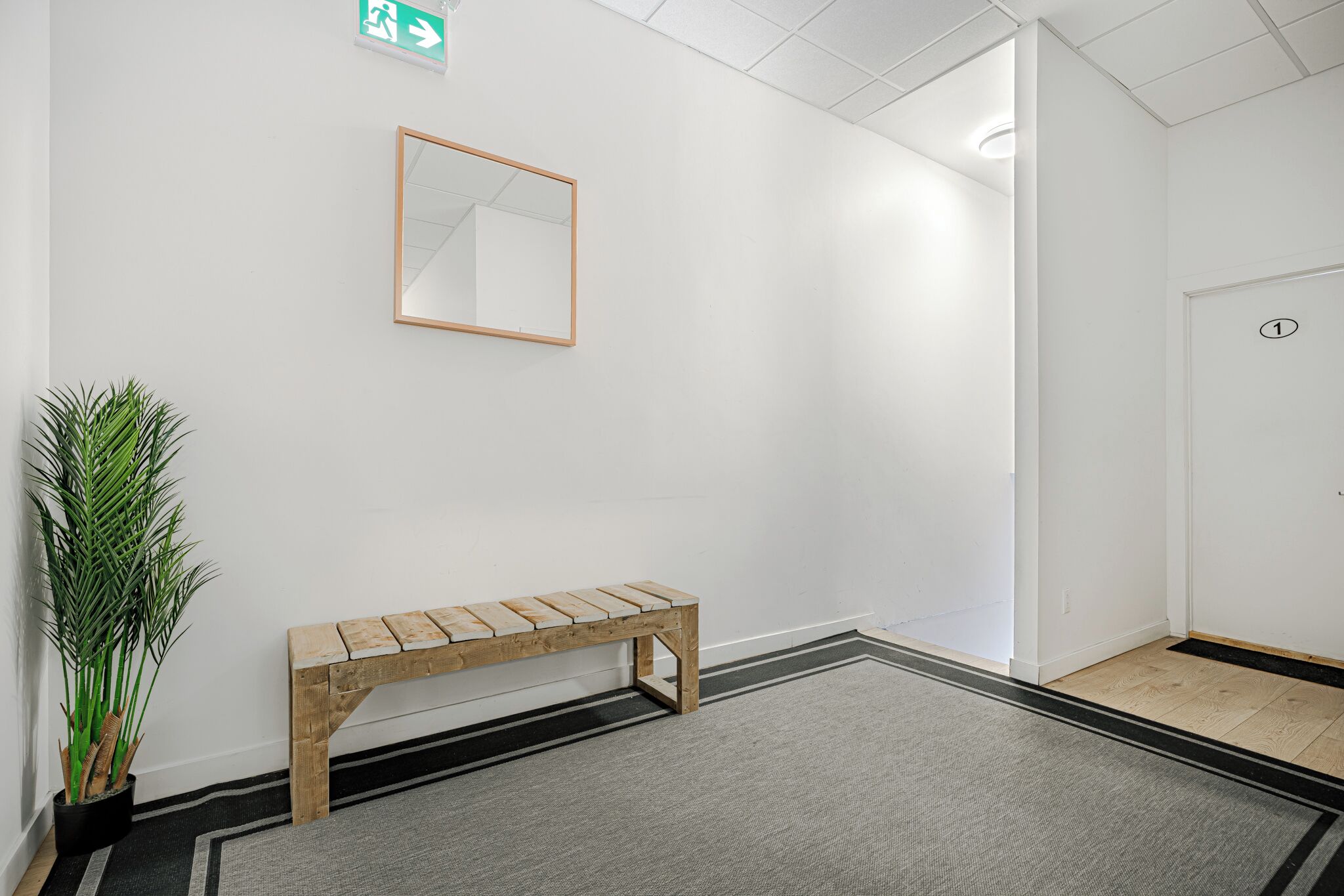 Un couloir épuré et minimaliste, avec un banc en bois, une plante en pot, un miroir et une porte portant le numéro 1.