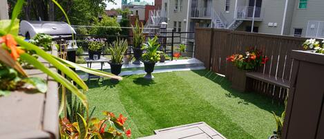 A backyard with artificial grass, potted plants, a barbecue grill, and a wooden deck.