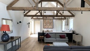 A spacious living area with a high ceiling, exposed wooden beams, a grey sofa, a small table with a lamp, and a kitchen in the background. - Stunning 19th century barn conversion in gorgeous Hampshire countryside (South Wonston)