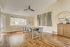 Un coin repas avec une table et des chaises en bois, un ventilateur de plafond, une grande fenêtre et un mur orné d’œuvres d’art abstraites.