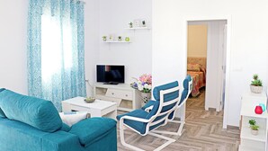 Ein Wohnzimmer mit einer Couch, einem Fernseher, einem kleinen Tisch, einem Sessel und einer Tür zu einem anderen Raum.