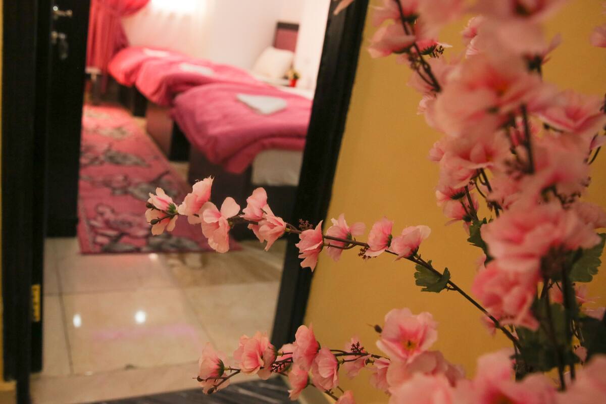 A room with a bed, a mirror reflecting the room, and pink flowers in the foreground.