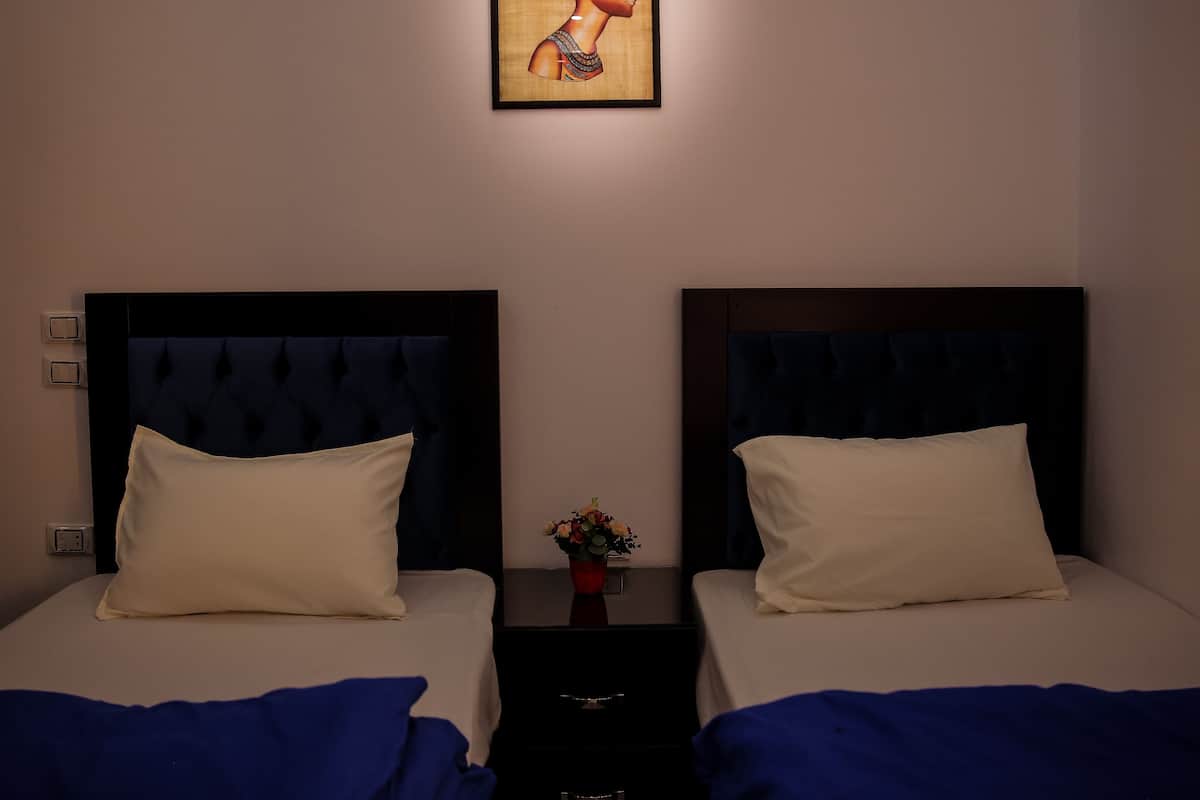 A hotel room with two beds, each with a dark headboard and white pillows. A small table with a vase of flowers is between the beds.