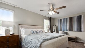 A bedroom with a bed, a nightstand, a dresser, a ceiling fan, a window with blinds, and a wall with a painting. - The Lake View: Family and business travel (Lehi)