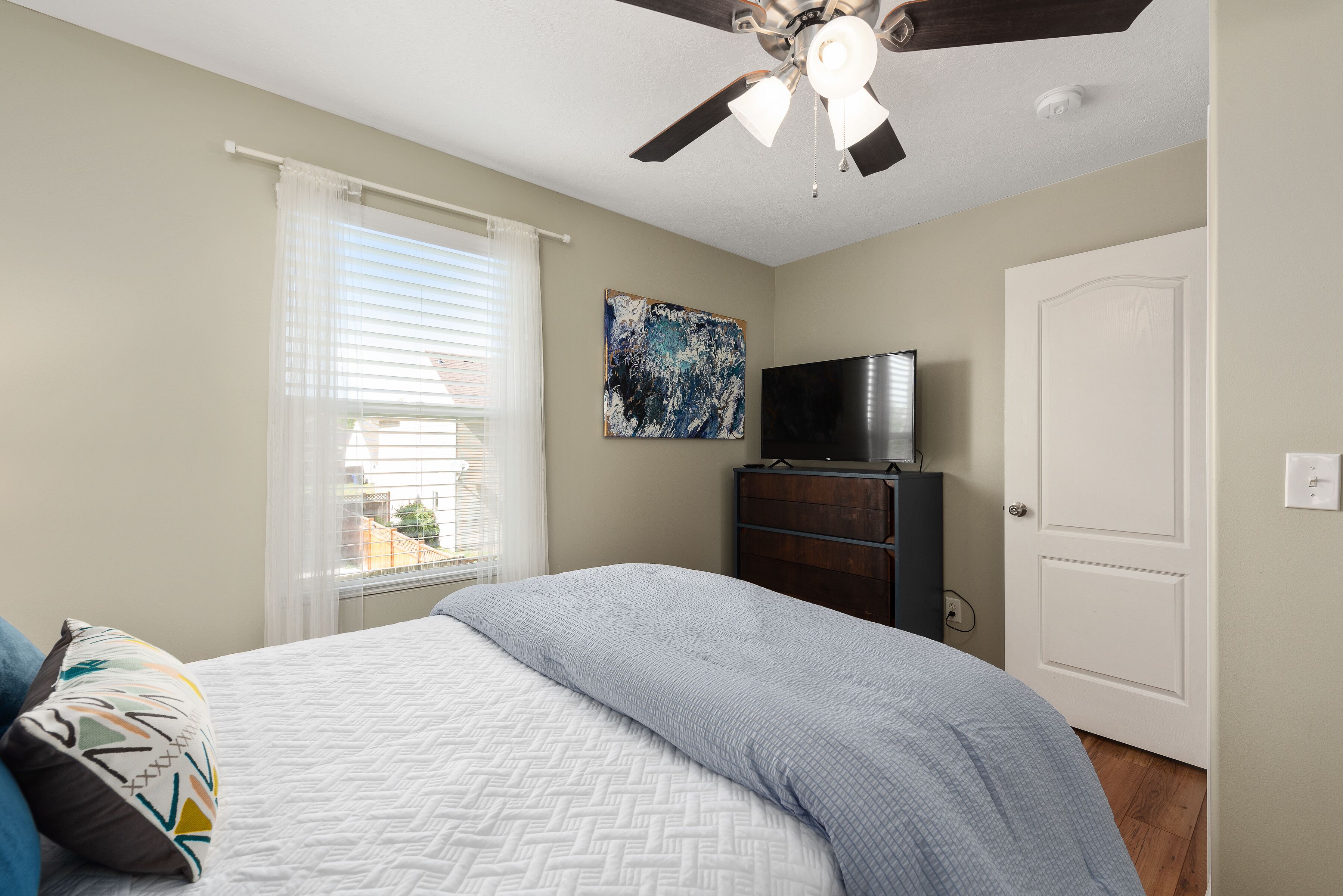 A bedroom with a bed, a ceiling fan, a TV, a dresser, and a door.