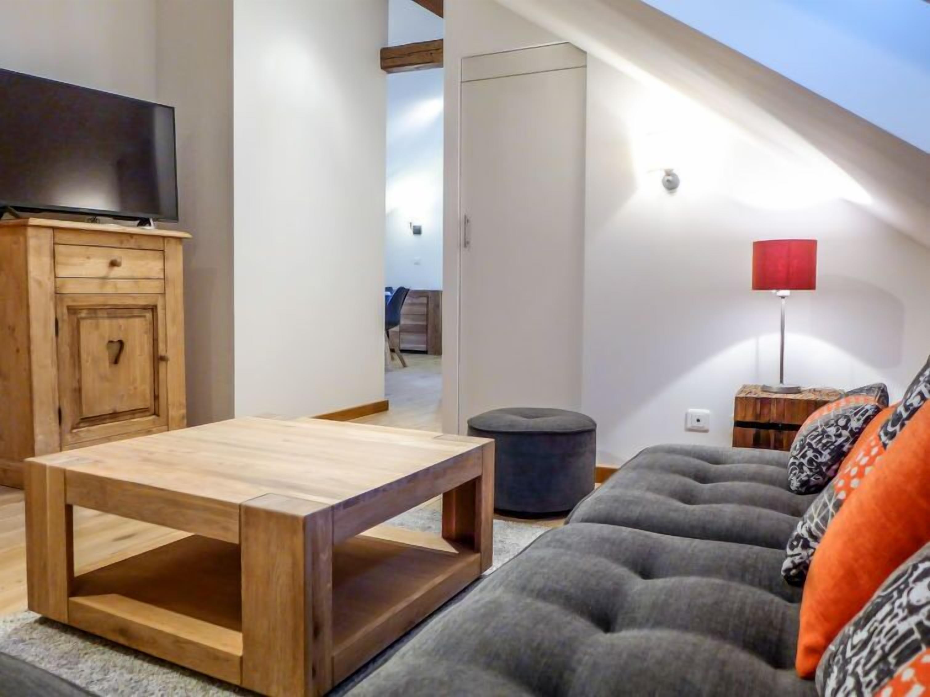 A modern living room with a wooden coffee table, grey sofa with patterned cushions, a red floor lamp, and a TV on a wooden cabinet.