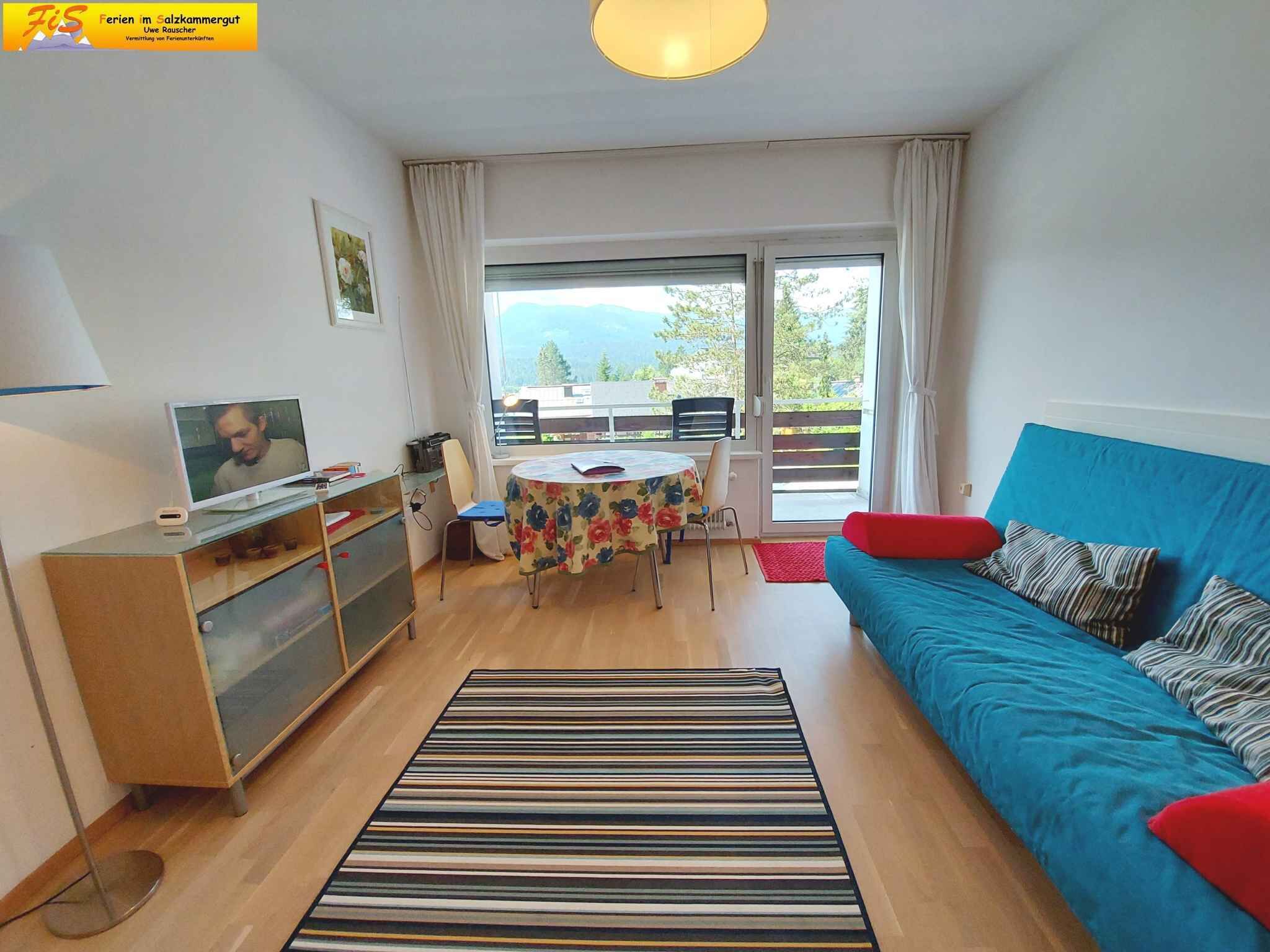 A modern living room with a blue sofa, a television, a dining table with chairs, and a large window with a view of greenery.