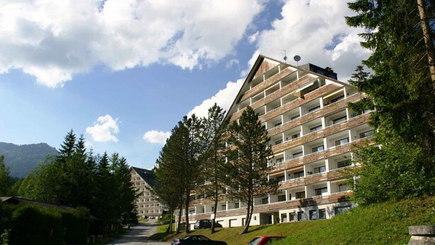 Un hôtel à plusieurs étages, doté d’une architecture moderne, avec des balcons et une vue sur la verdure et les montagnes environnantes.