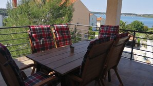 Ein Balkon mit Holztisch und -stühlen, roten und weißen Karokissen und Blick auf das Meer und die Gebäude.