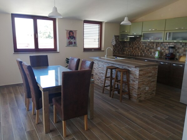 Una cocina moderna con una isla central, mesa de comedor y sillas. Hay ventanas con persianas, una lámpara colgante y armarios verdes.