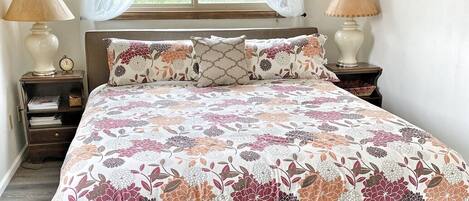 A bedroom with a floral bedspread, two bedside lamps, a clock, and a window with sheer curtains.