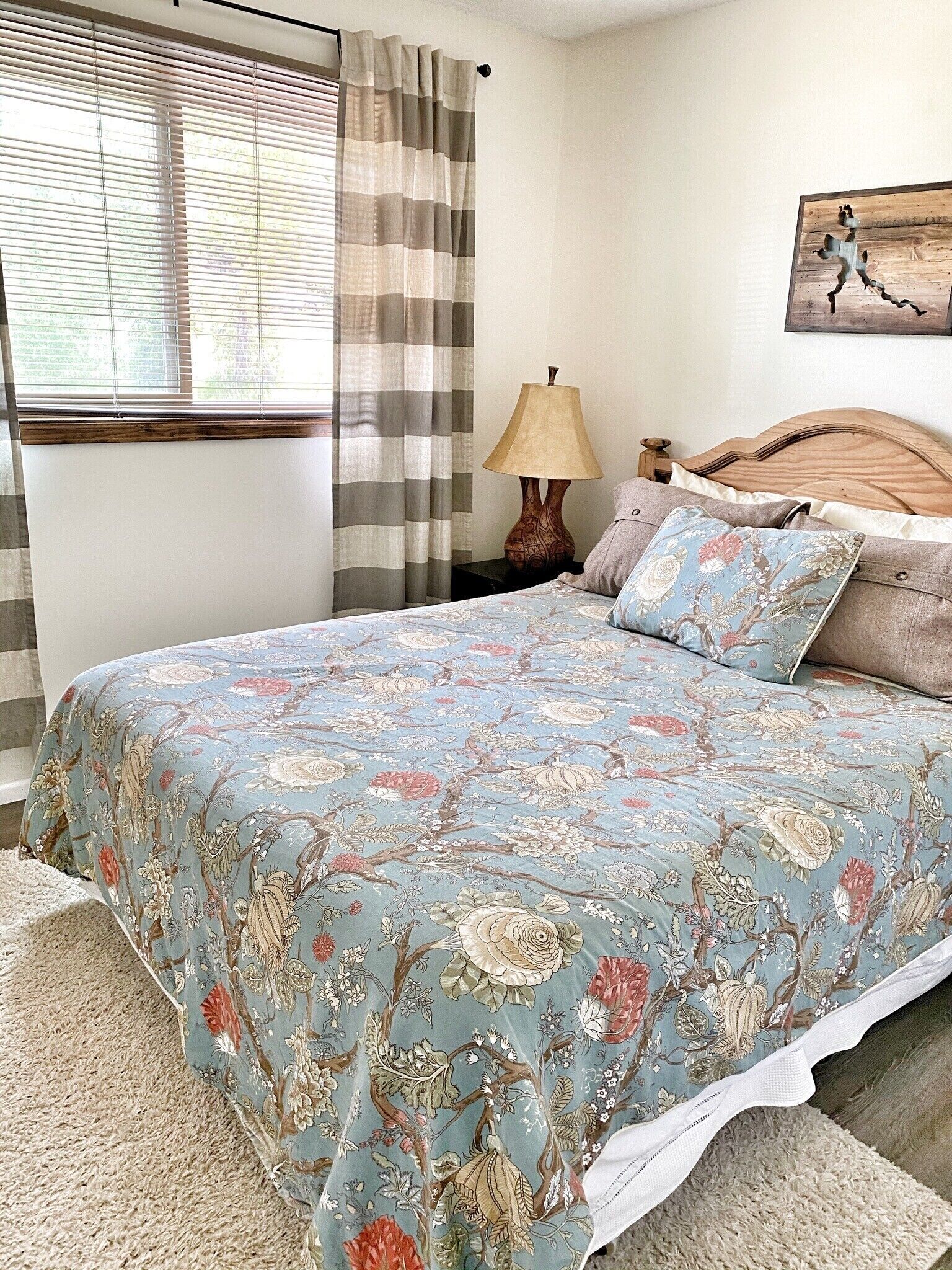 Un dormitorio con colcha estampada, cabecera de madera, mesita de noche con lámpara y ventana con persianas.
