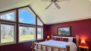Un dormitorio con una cama grande, un banco de madera, un ventilador de techo y varias ventanas que ofrecen vistas a un parque y montañas.