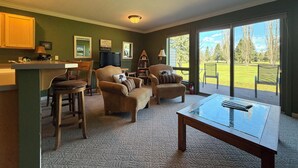 Sala de estar con una mesa de centro de vidrio, dos sillones, un sofá, un televisor y vistas a un área verde a través de amplios ventanales.
