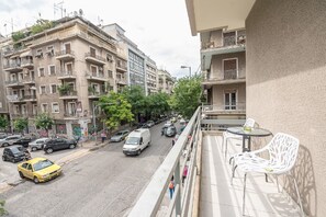 Blick von einem Balkon auf eine Straße mit einem mehrstöckigen Gebäude, Balkonen, parkenden Autos und einem gelben Taxi.
