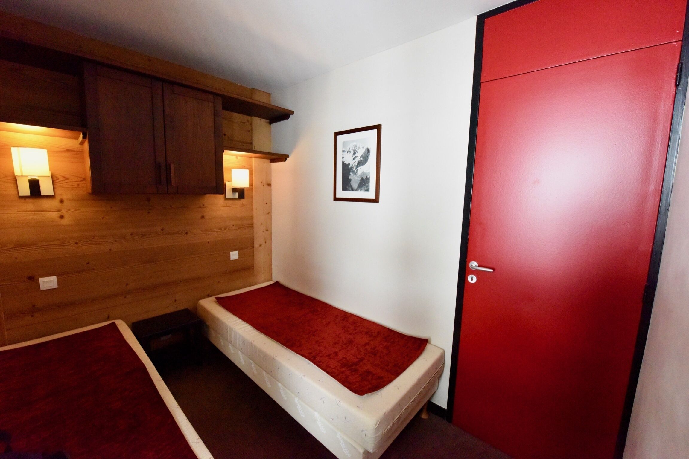 Ein kleines Zimmer mit einem Einzelbett, einer roten Tür, Holzvertäfelung und zwei Wandleuchten.