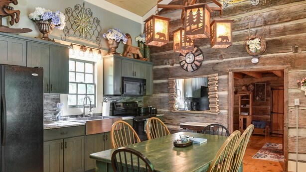 A rustic kitchen with wooden cabinets, a dining table with chairs, and decorative wall sconces. - Historic Log Cabin, Yard, Firepit, Fireplace, WIFI , Private (Maggie Valley)