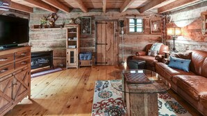 A rustic cabin interior with a wooden ceiling, a television, a fireplace, a leather sofa, and a coffee table. - Historic Log Cabin, Yard, Firepit, Fireplace, WIFI , Private (Maggie Valley)