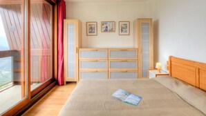 Ein Schlafzimmer mit großem Fenster, einem Bett, einem Nachttisch mit Lampe und einem Holzschrank.