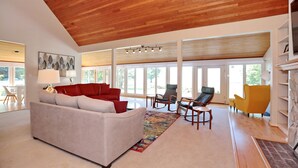 A spacious living room with a high wooden ceiling, a grey sofa with red cushions, and a colorful rug.