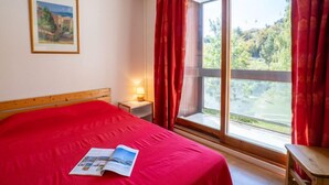 Quarto com uma cama grande, mesa de cabeceira com abajur, janela com cortinas vermelhas e um quadro na parede.