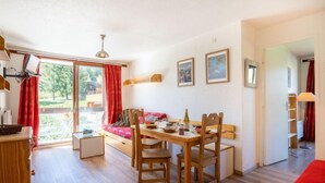 Uma sala de jantar com mesa e cadeiras de madeira, um sofá vermelho e vista para um terraço exterior através de uma porta de vidro.